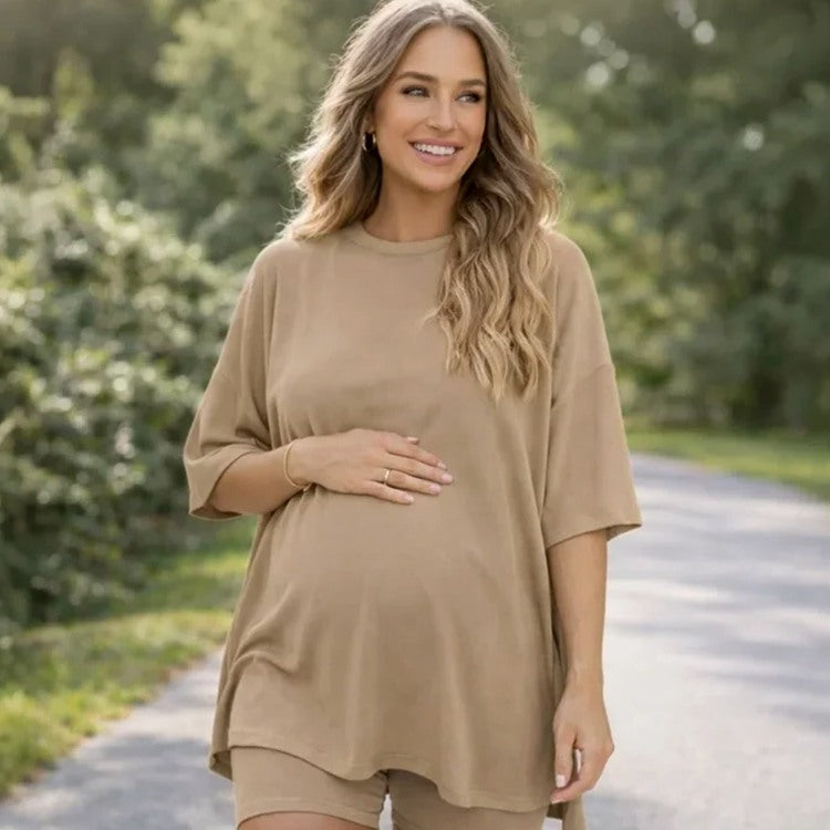 A pregnant woman wearing an oversized beige maternity t-shirt and matching shorts, standing on a paved path outdoors with a blurred green forest background.