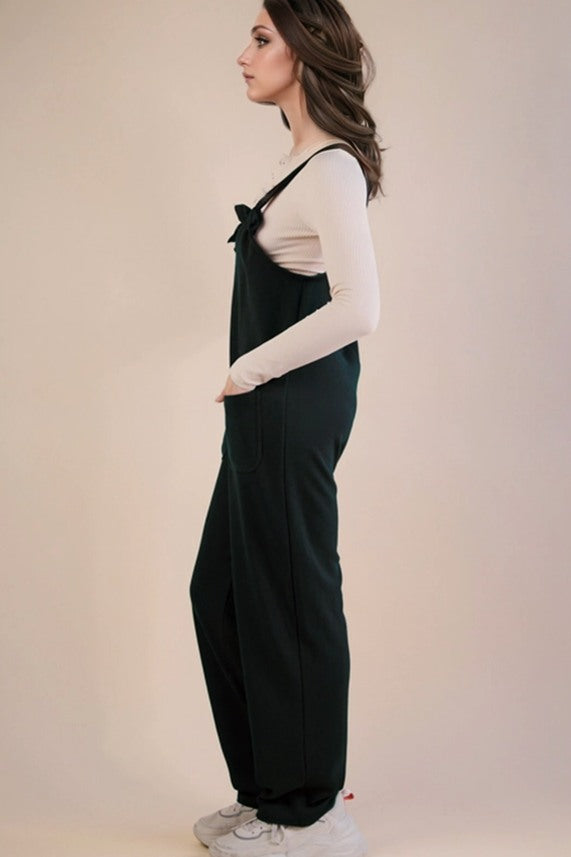A woman in profile wearing dark green wide-leg overall jumpsuit over a white long-sleeve top and white sneakers, standing against a plain beige studio background.