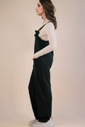 A woman in profile wearing dark green wide-leg overall jumpsuit over a white long-sleeve top and white sneakers, standing against a plain beige studio background.