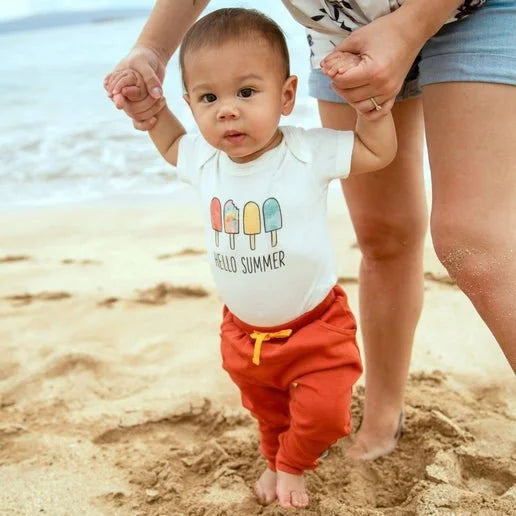 A baby walking on a sandy beach holding an adult's hand, wearing a white short-sleeve bodysuit with a colorful popsicle graphic and the words 'HELLO SUMMER' on the front, paired with bright orange pants.